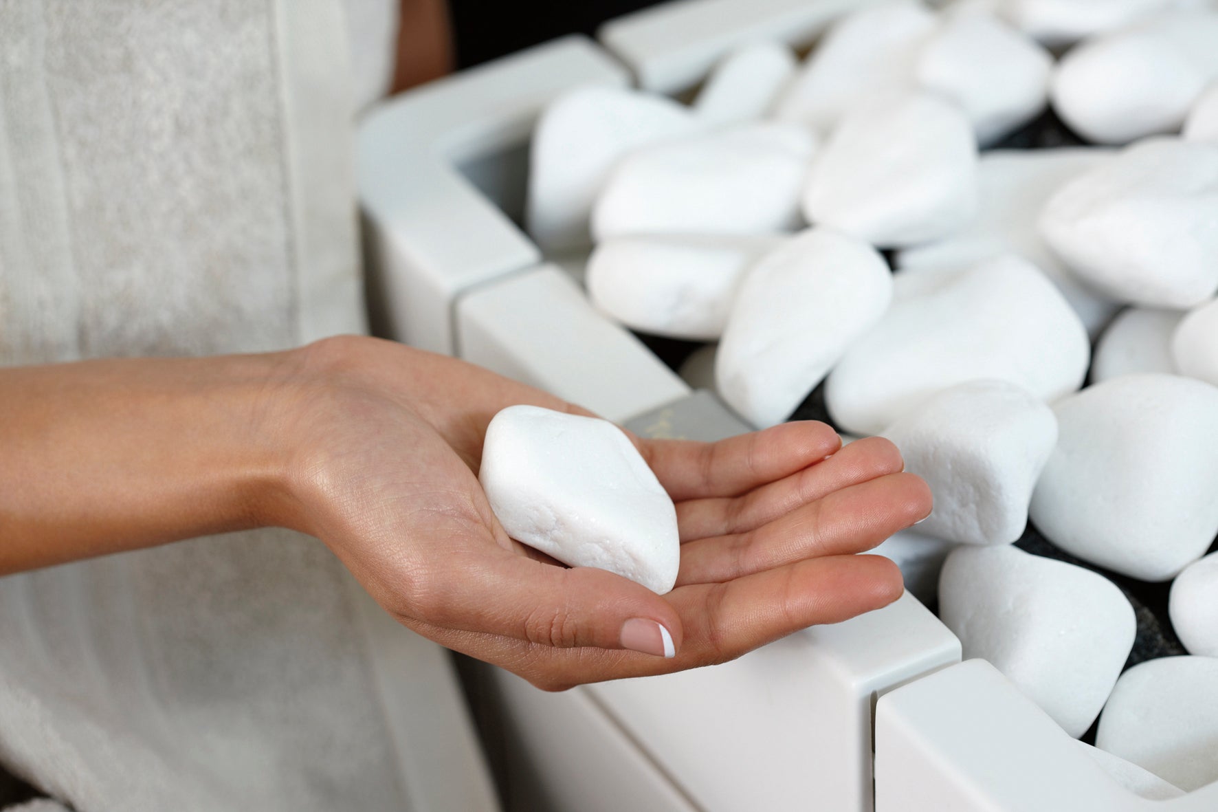 Woman's hand holding a single white decorative sauna stone with a collection of similar stones in the background