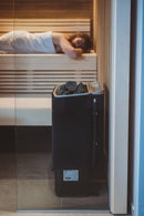 Side view of Tylo sense combi pure sauna heater with woman relaxing on sauna bench in background