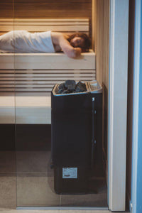 Side view of Tylo sense combi pure sauna heater with woman relaxing on sauna bench in background