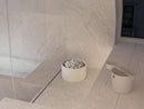 A minimalist white cylindrical sauna heater filled with small white pebbles sits on a light marble surface in a modern bathroom setting, alongside a matching white scoop or cup.