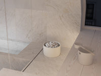 A minimalist white cylindrical sauna heater filled with small white pebbles sits on a light marble surface in a modern bathroom setting, alongside a matching white scoop or cup.