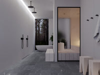 A modern minimalist bathroom interior with dark gray stone tile flooring, white walls, matte black fixtures, and a large black-framed mirror reflecting a spa-like treatment room. A freestanding oval bathtub is visible in the background near a window showing a forest view and a white cylindrical sauna heater in adjacent room