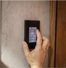 A hand is shown interacting with a slim, black digital Tuliki touchscreen sauna control panel mounted on a textured beige wall next to a wooden door.