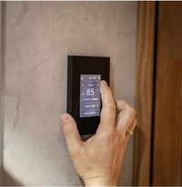 A hand is shown interacting with a slim, black digital Tuliki touchscreen sauna control panel mounted on a textured beige wall next to a wooden door.