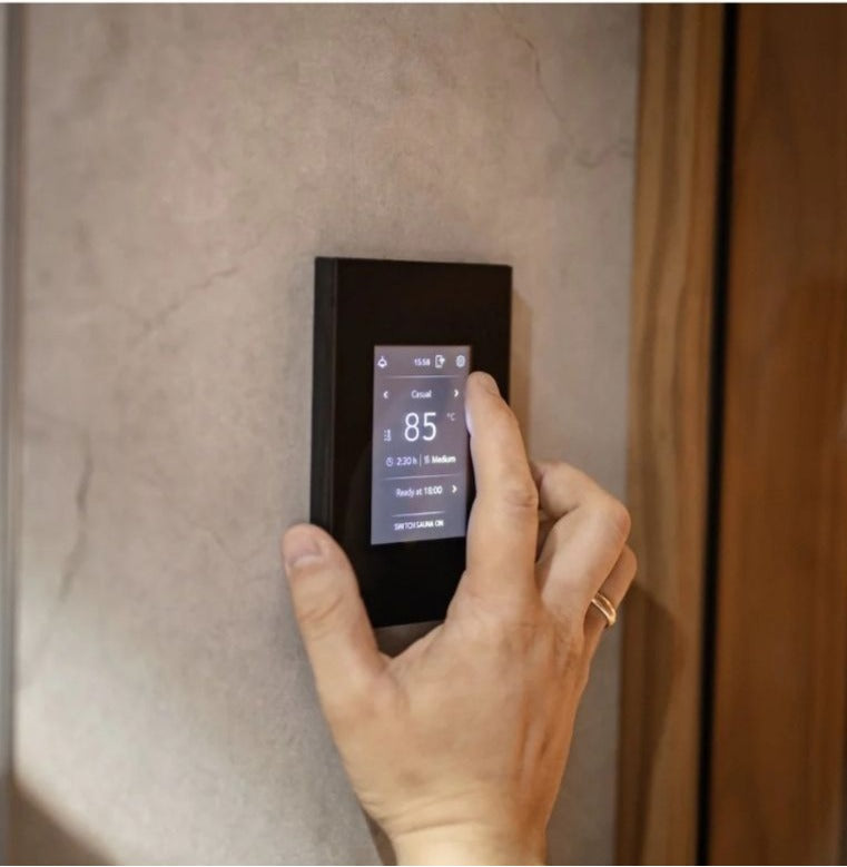 A hand is shown interacting with a slim, black digital Tuliki touchscreen sauna control panel mounted on a textured beige wall next to a wooden door.