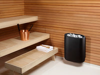 Sauna interior with wooden benches, a black sauna heater, and a copper bucket.