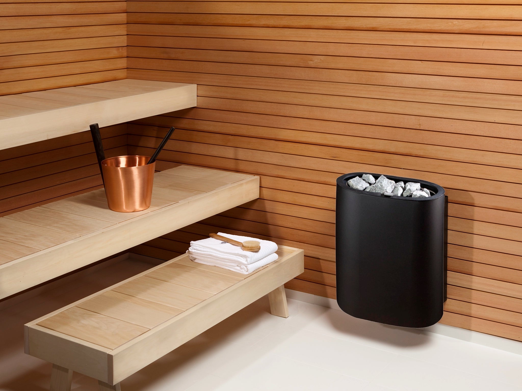 Sauna interior with wooden benches, a black sauna heater, and a copper bucket.